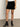 A person is wearing the quilted velvet mini skirt in black by Sanctuary Clothing, paired with black socks and heeled shoes, standing on a gray floor against a plain light background. Only their lower half is visible.