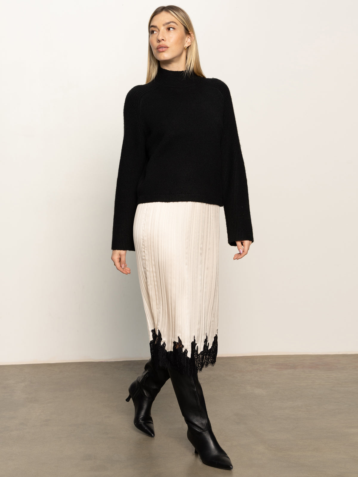 A woman in a black long-sleeve sweater, black knee-high boots, and the Sanctuary Clothing evermore pleated midi skirt in light oat stands against a plain off-white wall and gray floor.