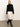 A woman in a black long-sleeve sweater, black knee-high boots, and the Sanctuary Clothing evermore pleated midi skirt in light oat stands against a plain off-white wall and gray floor.