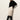 A woman with straight light brown hair and a neutral expression stands against a white wall wearing Sanctuary Clothing's evermore pleated midi skirt in light oat, styled with a black turtleneck sweater and black knee-high heeled boots.