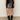 A person is seen from the waist down wearing the Sanctuary Clothing broadway leather skirt in black, paired with black patent boots and a red top, standing against a plain light-colored wall.