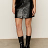 A person is seen from the waist down wearing the Sanctuary Clothing broadway leather skirt in black, paired with black patent boots and a red top, standing against a plain light-colored wall.