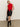A woman poses against a plain background wearing Sanctuary Clothing's broadway leather skirt in black, paired with a sleeveless red neck-tie top, black heeled boots, and holding a black studded handbag. One hand rests in her pocket.