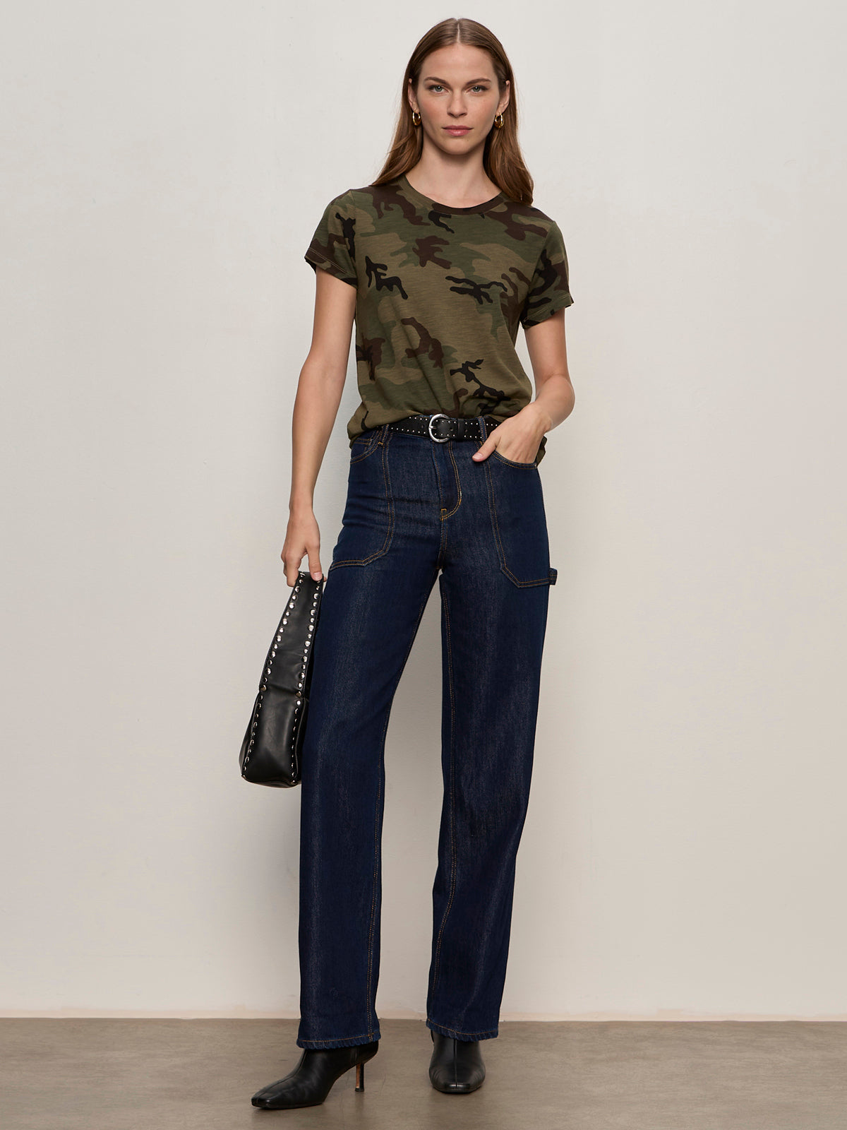 A woman with long hair and minimal jewelry stands against a plain background, wearing Sanctuary Clothing's the perfect tee hiker camo, high-waisted dark blue jeans, black heeled boots, and holding a black studded handbag.