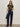A woman with long hair and minimal jewelry stands against a plain background, wearing Sanctuary Clothing's the perfect tee hiker camo, high-waisted dark blue jeans, black heeled boots, and holding a black studded handbag.