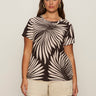 A woman wears Sanctuary Clothing's "the perfect tee mesa palm extended sizing," a short-sleeve beige top with dark brown palm leaves, paired with light beige shorts, standing against a plain, light background.