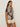 A woman with long brown hair wears Sanctuary Clothing's the perfect tee mesa palm extended sizing—a short-sleeved black and beige patterned top—paired with light beige shorts, posing confidently against an off-white background.