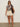 A woman in Sanctuary Clothing's the perfect tee mesa palm (extended sizing), cream shorts, and slide sandals stands against a plain wall, wearing sunglasses, carrying a dark brown shoulder bag, and wearing her hair down.