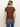 A woman with blonde hair in a low ponytail is seen from behind, wearing the perfect tee boheme by Sanctuary Clothing�a brown short-sleeve top with a subtle floral pattern�paired with blue jeans against a plain light background.