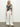 Dressed in Sanctuary Clothing's perfect tee inked impressions, a person with long blonde hair stands against a plain background. They pair the black and white patterned shirt with white wide-leg pants and black sandals, gazing at the camera in a relaxed posture.