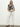Sporting the Sanctuary Clothing's Perfect Tee in Inked Impressions, a long-haired individual pairs it with white wide-leg pants and black sandals. They stand against a plain white wall, hands clasped in front.