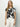 A woman with long blonde hair models Sanctuary Clothing's "the perfect tee inked impressions," featuring a black and white floral pattern, paired with white pants. She stands against a plain white backdrop, accessorized with gold jewelry.