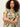 A woman with curly hair stands confidently with arms crossed, modeling the Sanctuary Clothing “the perfect tee palm,” a short-sleeve shirt featuring a green and pink tropical leaf pattern, against a plain background.
