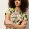 A woman with curly hair stands confidently with arms crossed, modeling the Sanctuary Clothing “the perfect tee palm,” a short-sleeve shirt featuring a green and pink tropical leaf pattern, against a plain background.
