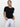 A person with long dark hair tied back stands against a plain background, wearing the perfect tee remix black from Sanctuary Clothing and white pants. They gaze at the camera neutrally, hands resting in their pockets.