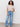 A woman with curly hair stands against a plain wall, wearing the Sanctuary Clothing perfect tee remix vapor rose, blue wide-leg jeans, and light-colored shoes. She has one hand in her pocket and smiles softly.