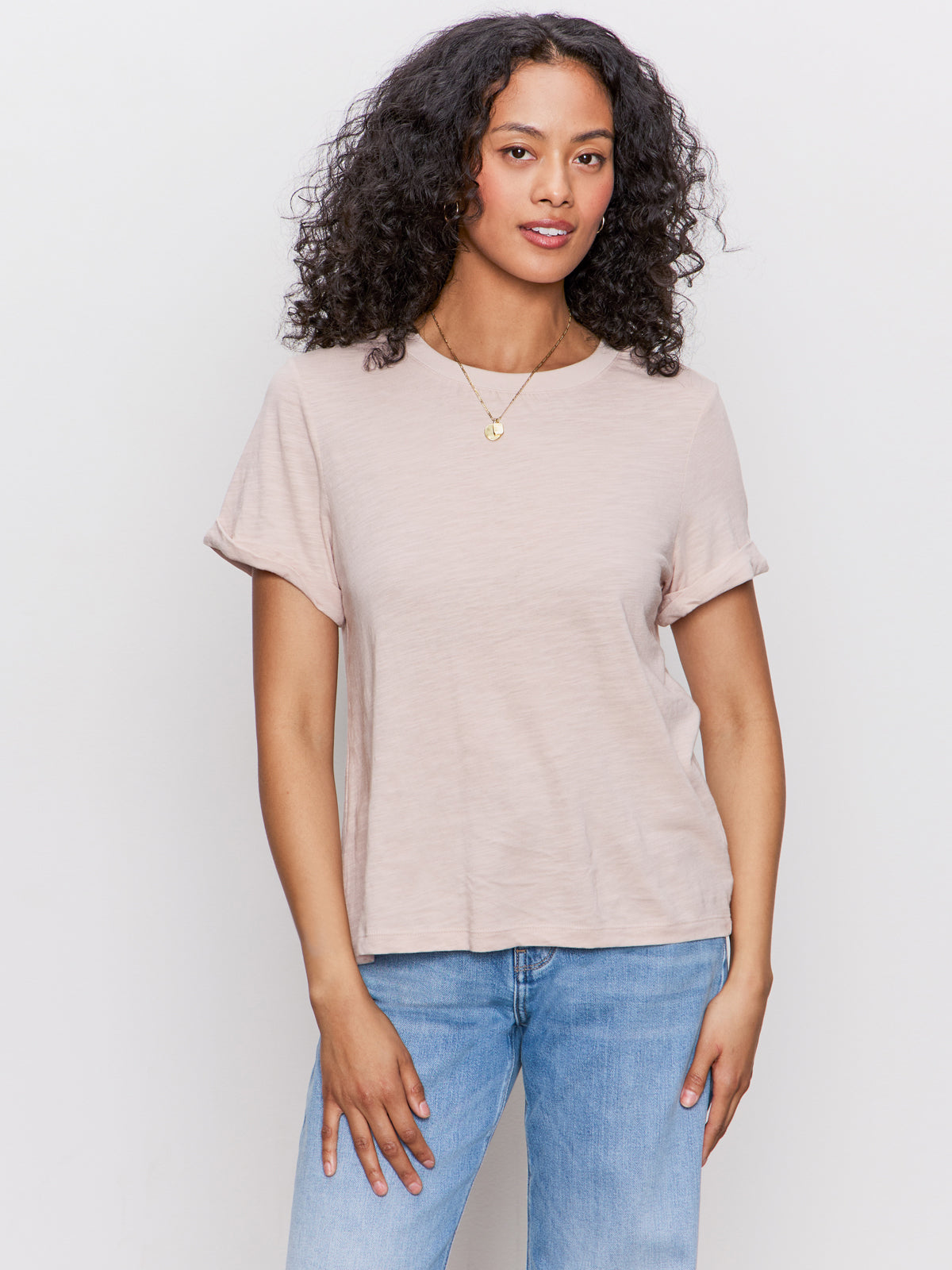 Woman with curly hair wearing the Sanctuary Clothing perfect tee remix in vapor rose, paired with blue jeans and a gold necklace, standing against a plain light background and smiling softly.