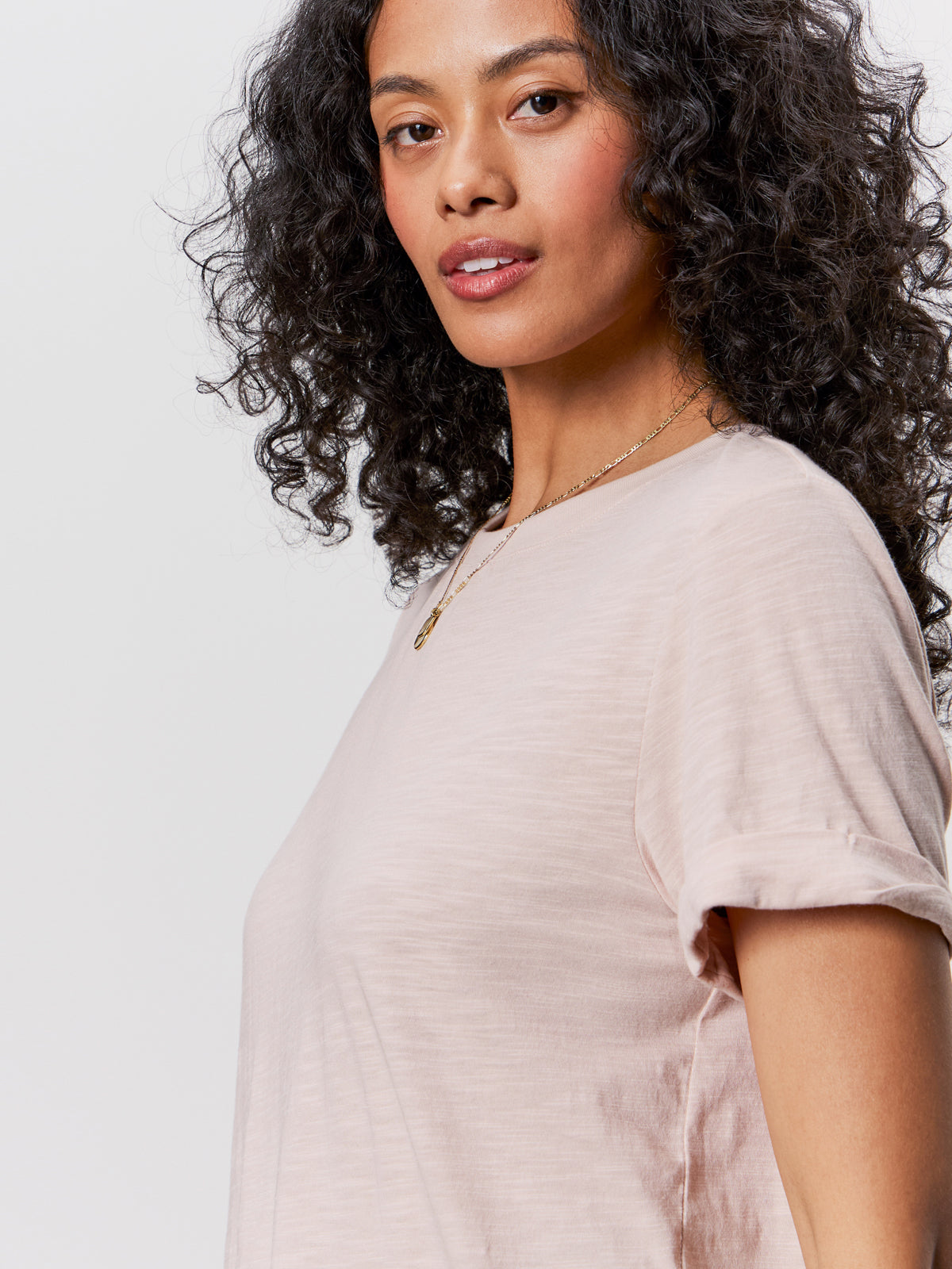 A woman with curly dark hair wears the Sanctuary Clothing perfect tee remix vapor rose, paired with a gold necklace, as she poses confidently against a plain light background.
