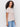 A woman with curly hair models the Sanctuary Clothing perfect tee remix vapor rose, a light pink short-sleeve t-shirt, paired with blue jeans. She stands sideways against a plain background, gazing at the camera with a neutral expression.