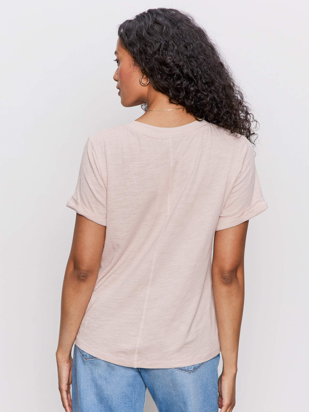 A woman with curly hair faces away, wearing the Sanctuary Clothing perfect tee remix in vapor rose and light blue jeans, set against a plain white background.