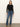 A woman with straight light brown hair wears the Sanctuary Clothing long sleeveperfect mesh tee in black, paired with high-waisted blue jeans, a black belt, and tan shoes. She stands against a plain light wall with a neutral expression.