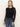 A woman with straight, light brown hair wears the Sanctuary Clothing long sleeveperfect mesh tee in black, styled with blue high-waisted jeans and a black belt with a gold buckle, standing against a plain light background.