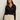 A woman with long brown hair wears the Sanctuary Clothing pucker up dreamgirl black textured button-up cardigan with a V-neck, paired with cream pants, standing against a plain off-white background and looking confidently at the camera.