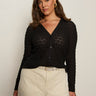 A woman with long brown hair wears the Sanctuary Clothing pucker up dreamgirl black textured button-up cardigan with a V-neck, paired with cream pants, standing against a plain off-white background and looking confidently at the camera.