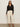 A woman stands against a light wall wearing the Sanctuary Clothing "pucker up dreamgirl black" textured button-up blouse, paired with light beige high-waisted pants, a beige belt, black heeled sandals, and gold hoop earrings.