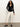 A woman with long curly hair wears the Sanctuary Clothing dreamgirl lace button up black top, paired with off-white ankle-length pants and black heeled mules. She stands before a plain white wall on a gray floor.