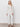 A woman with long, wavy blonde hair wears the Sanctuary Clothing dreamgirl lace button up in chalk, paired with white pants and black sandals. She stands against a plain white background with a relaxed expression.