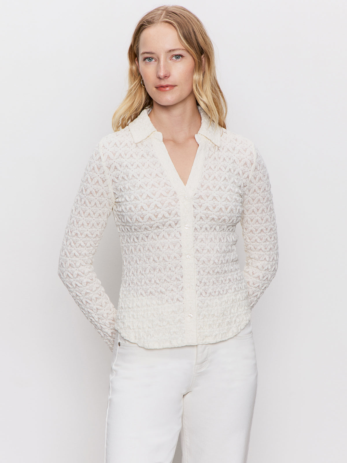 A blonde woman poses against a white background, wearing the Sanctuary Clothing dreamgirl lace button up chalk blouse and white pants, with her hands behind her back and looking at the camera.