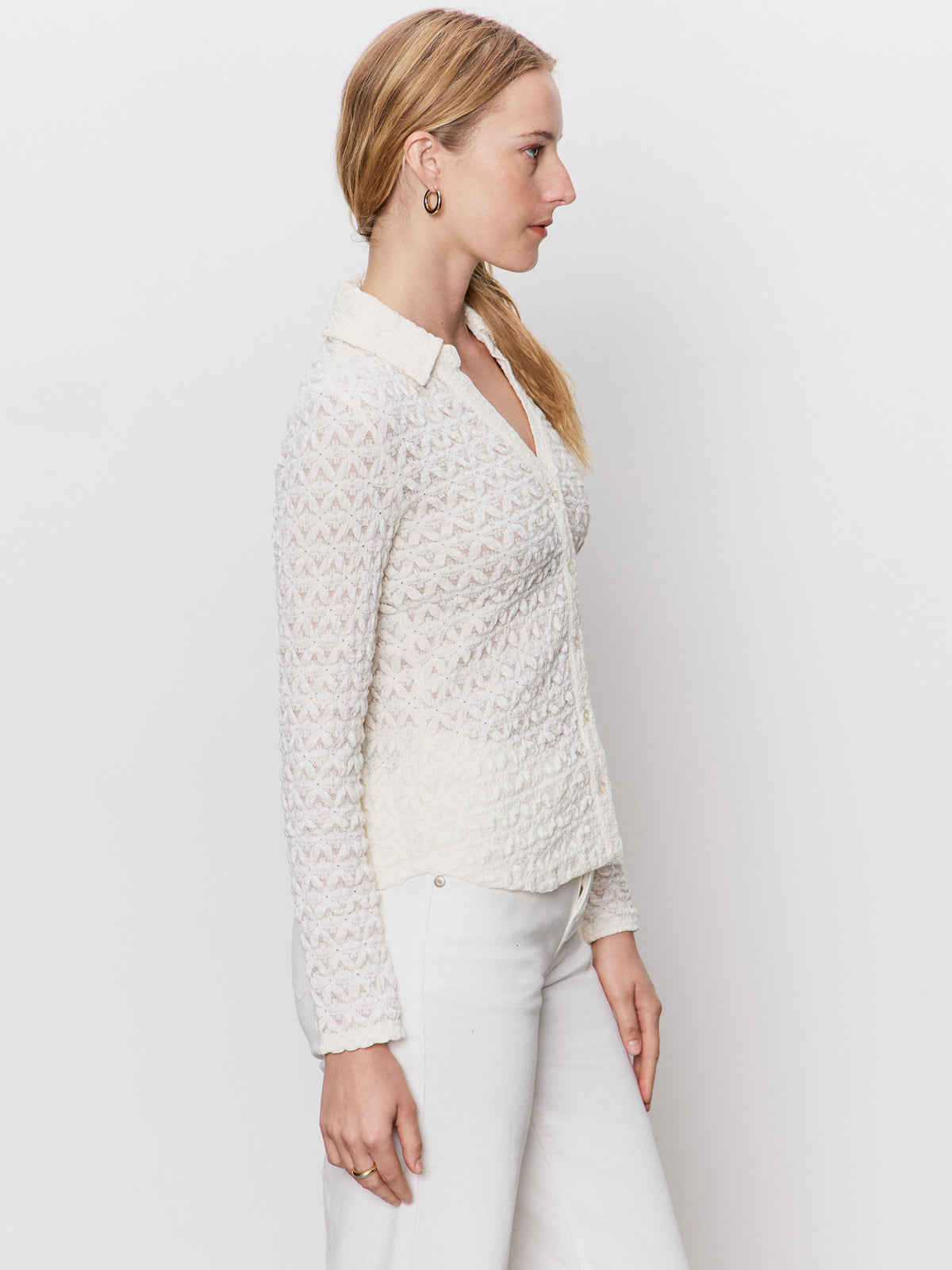 A woman with light skin and blonde hair in side profile wears the Sanctuary Clothing dreamgirl lace button up chalk blouse with white pants, standing against a plain white background.