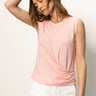 A woman in the Sanctuary Clothing draped tank cherry blossom and white pants stands against a plain background. One hand is on her head, the other relaxed, eyes closed, wearing a bracelet and necklace.