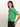 A person with long brown hair wears the Sanctuary Clothing "all day eyelet tee palm" alongside black pants. They face slightly right, sporting a neutral expression that highlights the detailed design of the green top.