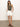 A woman in a ribbed racer tank kalamata by Sanctuary Clothing and white shorts stands against a plain backdrop. She's wearing brown sandals and a necklace, hands in her pockets, on a neutral indoor floor.