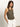 A woman with long brown hair is wearing a ribbed racer tank in kalamata by Sanctuary Clothing and white shorts, her hands in pockets, standing against a plain background and wearing a necklace.