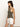 A woman with long brown hair is wearing a ribbed racer tank in kalamata by Sanctuary Clothing and white shorts. She faces sideways against a plain white background, accessorized with a gold necklace and rings.