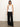 A person with long hair stands against a plain backdrop wearing a Sanctuary Clothing ribbed racer tank in white, along with loose black pants and sandals. They accessorize with a simple necklace featuring a round pendant, adopting a relaxed posture.
