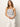 A person with long hair models the ribbed racer tank in white black stripe by Sanctuary Clothing, paired with white pants, standing against a plain background. They clasp their hands and look directly at the camera.