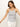 A woman in a Sanctuary Clothing ribbed racer tank with black stripes and white pants stands on a plain white background, arms slightly open. She has long brown hair, a gold necklace, and hoop earrings.