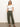 A person confidently poses with hands in pockets, dressed in Sanctuary Clothing's off shoulder rib tee white and olive green cargo pants, paired with brown sandals. The backdrop is a plain light color, and the floor is smooth gray.