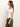 A woman with long brown hair wearing a Sanctuary Clothing off-shoulder rib tee in white and olive green pants stands against a plain background, looking to the left with her arms relaxed by her sides.