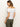 A brown-haired woman in a white Sanctuary Clothing off-shoulder rib tee and green pants looks down to her side against a plain, light background.