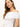 A woman with long brown hair is wearing Sanctuary Clothing's off shoulder rib tee in white, paired with gold hoop earrings and a matching necklace. She stands against a plain light background, looking towards the camera with a slight smile.