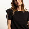 A person with long brown hair is wearing a black relaxed muscle tee by Sanctuary Clothing. They are positioned in front of a neutral background, gazing slightly to the side.