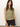 A woman with long brown hair wears the Sanctuary Clothing ribbed ruched tank in sage and black shorts, standing before a plain background. Exuding confidence, she accessorizes with a gold necklace and ring, her hands relaxed by her sides.