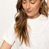 A woman with long, wavy hair wears Sanctuary Clothing's embroidered perfect tee in white. She smiles slightly downward, touching her ear to showcase gold hoop earrings against a light, neutral background.