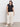 A person with long dark hair, wearing Sanctuary Clothing's knotted muscle tee black extended sizing, cream wide-leg pants, and white sneakers stands against a plain backdrop. They look towards the camera with a neutral expression.