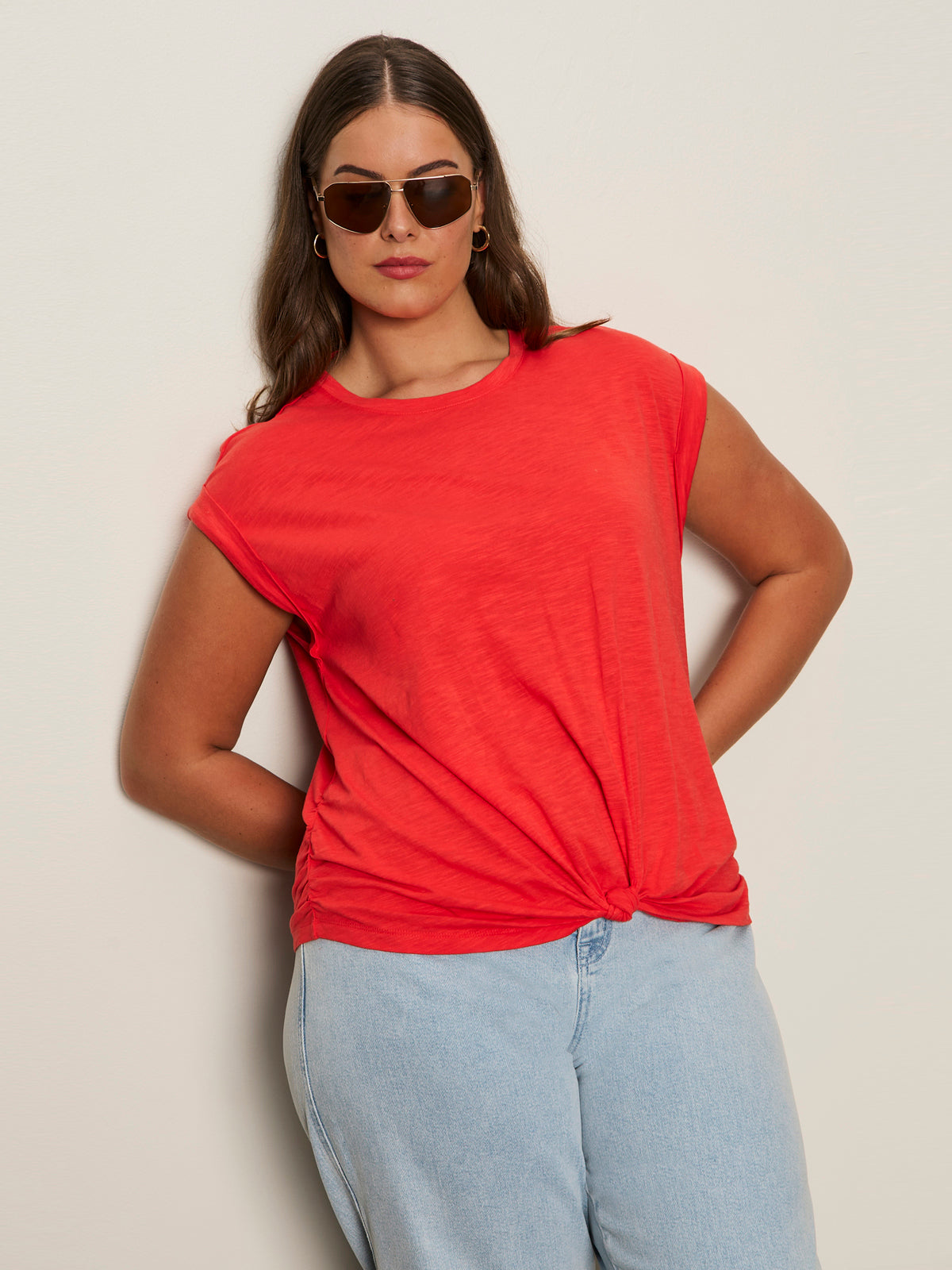 A woman wears the Sanctuary Clothing knotted muscle tee in cayenne (extended sizing) with light blue jeans. She stands against a light wall, has long brown hair, sunglasses, and her hands are behind her back.